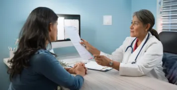 A female healthcare professional in a white coat holding a medical chart while discussing test results with a female patient in a medical office.