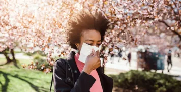Woman blowing nose outdoors surrounded by spring tree pollen and seasonal allergy triggers