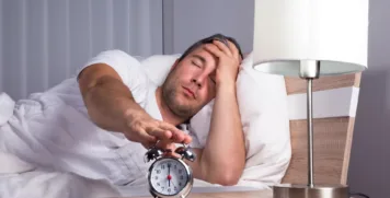 Exhausted man lying in bed holding head while reaching for alarm clock