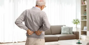 Senior man standing in living room holding lower back in discomfort from chronic back pain